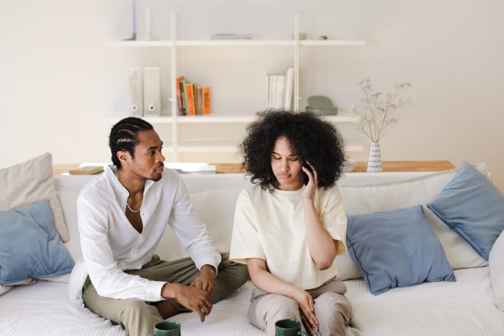 African American couple relaxing at home, sitting on sofa, talking and using phone.